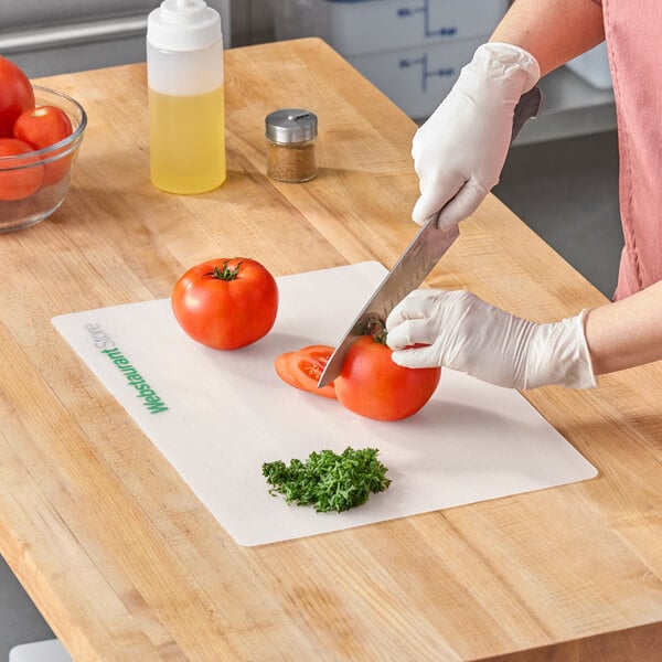 A flexible white cutting board mat with the WebstaurantStore logo, shown in use for slicing tomatoes and chopping herbs.