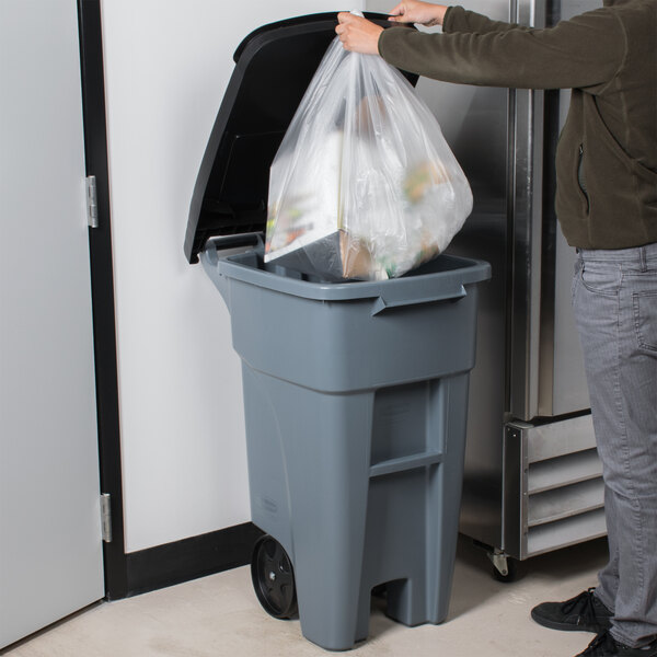 A man putting a plastic bag of garbage into a Rubbermaid wheeled trash can.