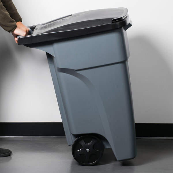 A man pushing a Rubbermaid grey wheeled rectangular trash can.