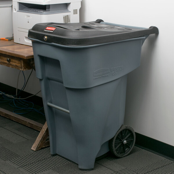 A grey Rubbermaid trash can with wheels and a locking lid next to a printer.