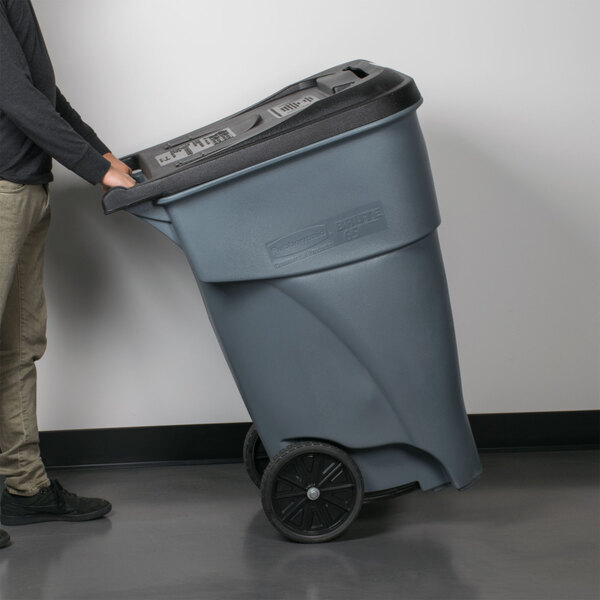 A man pushing a Rubbermaid 65 gallon grey rectangular trash can.