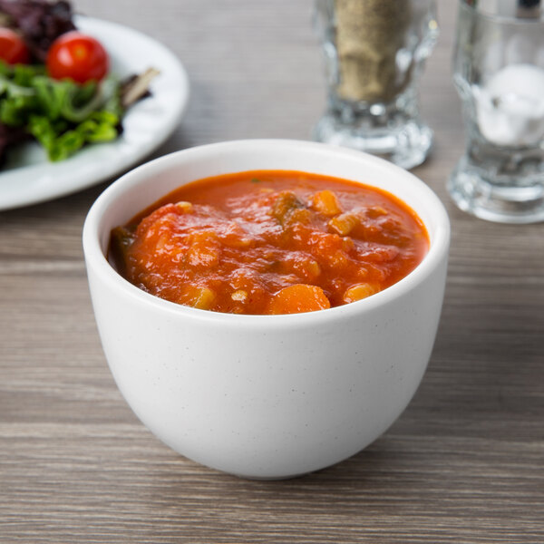 A white Libbey Driftwood satin matte porcelain bouillon bowl filled with red soup on a table with a plate of salad and a glass.