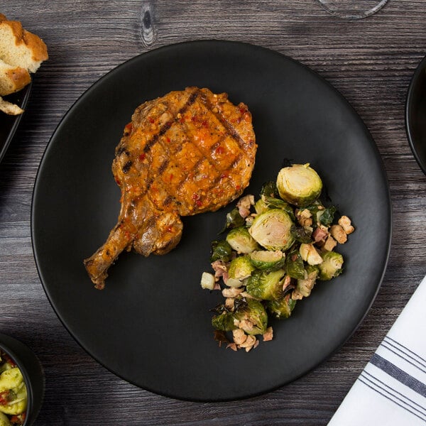 A Libbey Driftstone Onyx matte porcelain coupe plate with a pork chop and brussels sprouts on a table.