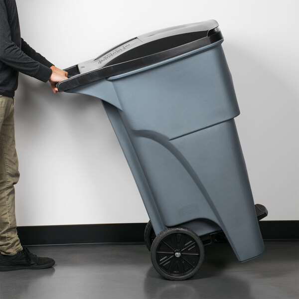 A person using a Rubbermaid 65 gallon wheeled trash can to push trash.