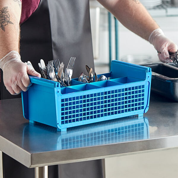 A person in a black apron placing a blue plastic container full of silverware into a blue Carlisle flatware rack.