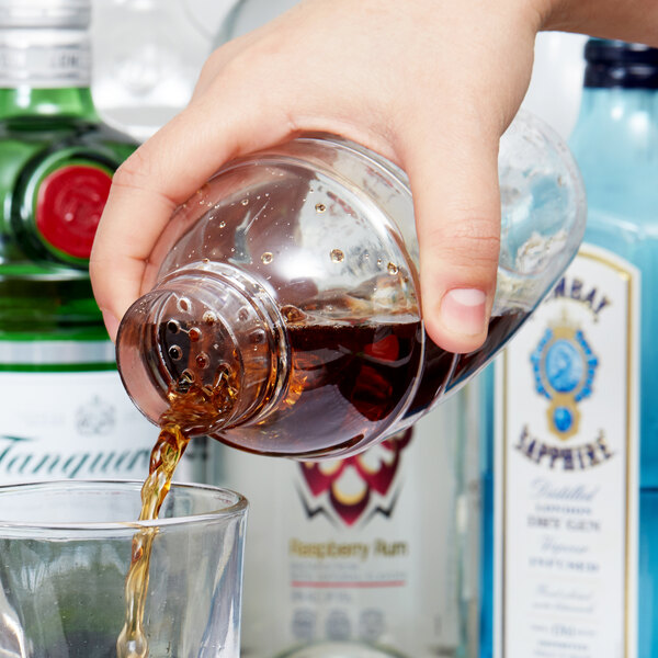 A hand pouring brown liquid from a bottle into a clear plastic shaker.