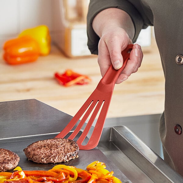 A hand holding a red Mercer Culinary Hell's Tools slotted spatula over a pan with a burger.