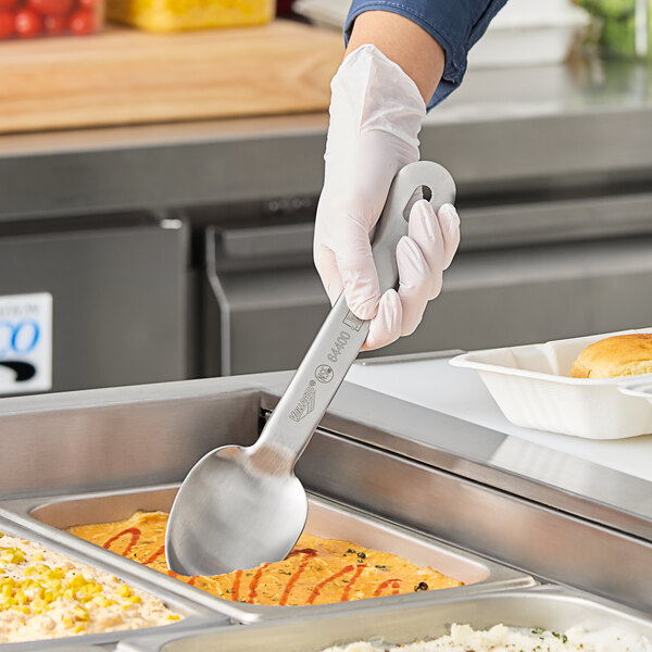 A person in gloves holding a Vollrath stainless steel serving spoon over a tray of food.