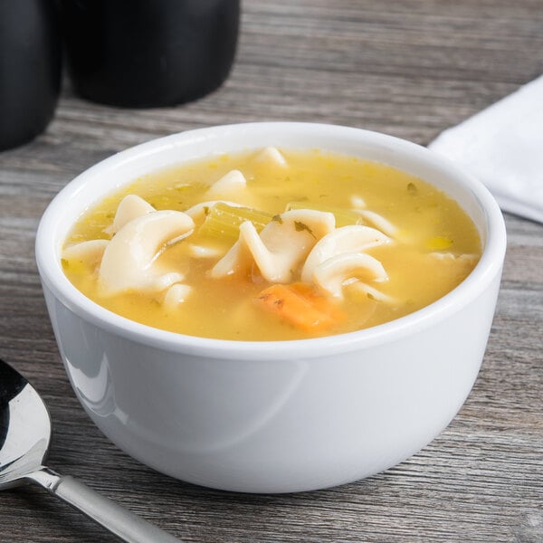 A white Elite Global Solutions round melamine bowl filled with soup, noodles, and vegetables with a spoon.