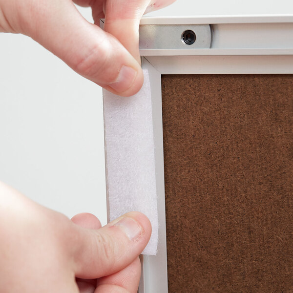 A person's hands using 3M white multi-purpose fasteners to hold a white piece of paper to a brown board.