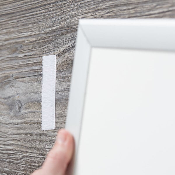 A hand holding a white frame next to a piece of white Scotch multi-purpose fastener tape.