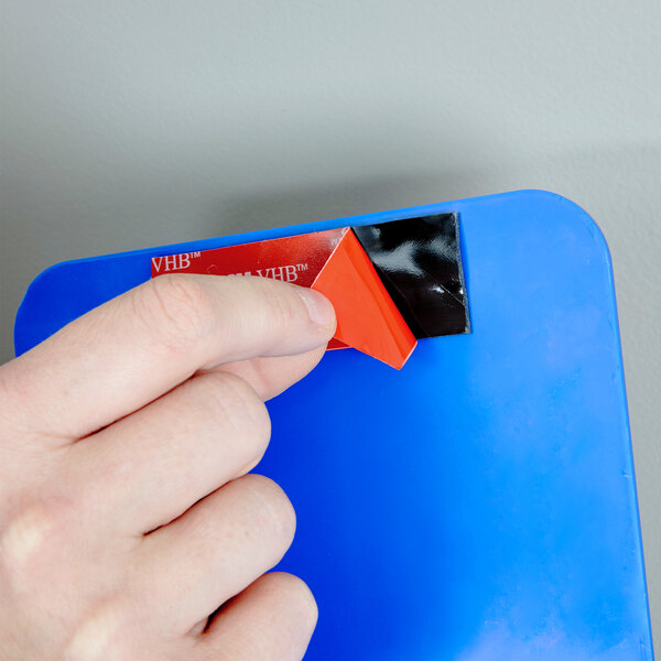 A hand using a red and black 3M Scotch Mounting Strip to hang a sign.