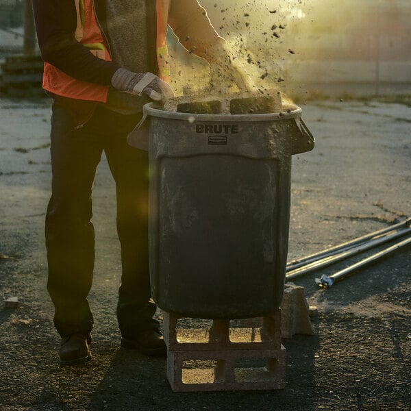 A man in an orange vest throwing debris into a Rubbermaid Brute 55 gallon gray trash can.