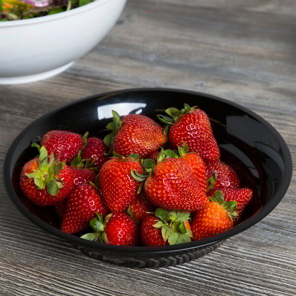 A black Fineline low profile plastic catering bowl filled with strawberries on a wooden table.