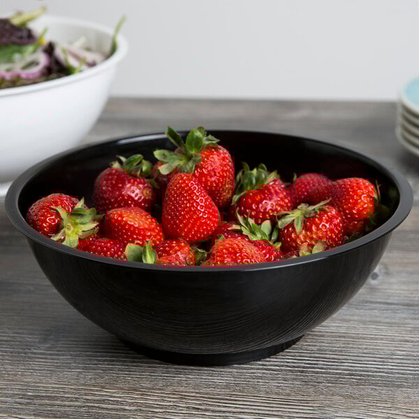 A black Fineline high profile plastic catering bowl filled with strawberries on a table with a bowl of salad.