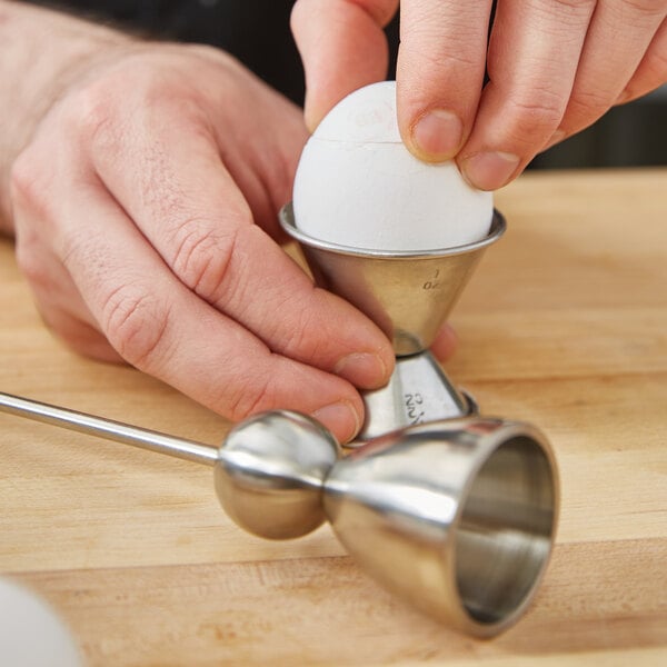 A hand using a Matfer Bourgeat stainless steel egg topper to crack an egg into a metal cup.