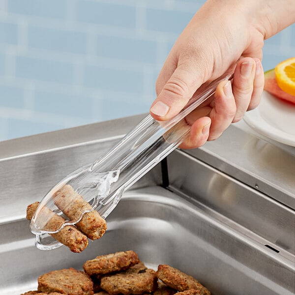 A hand using Carlisle clear utility tongs to serve food from a glass container.