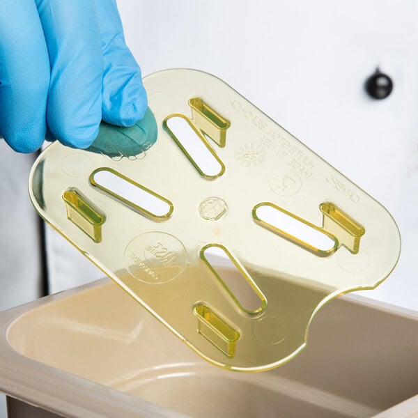 A hand in a blue glove holding a yellow plastic Cambro drain tray.