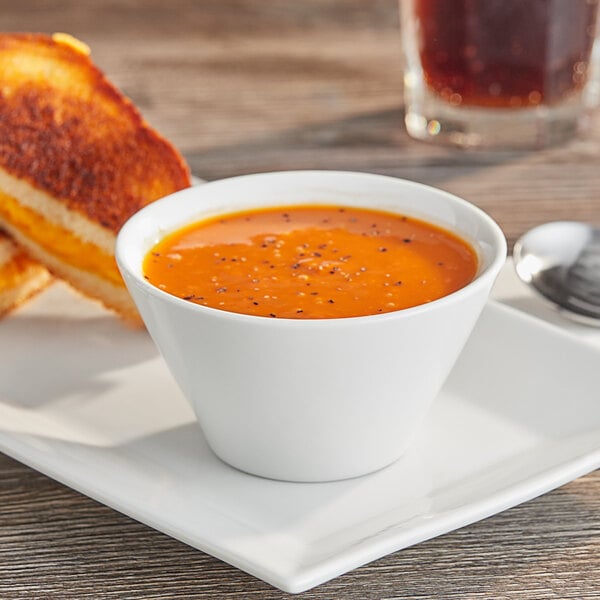 A bowl of Acopa bright white porcelain bouillon with soup and bread on a table with a grilled sandwich.