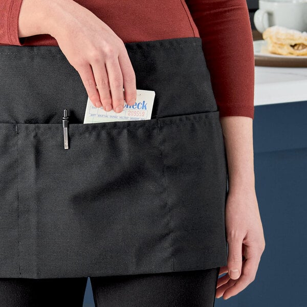 A person wearing a black waist apron with three pockets, placing a notepad into one of the pockets.