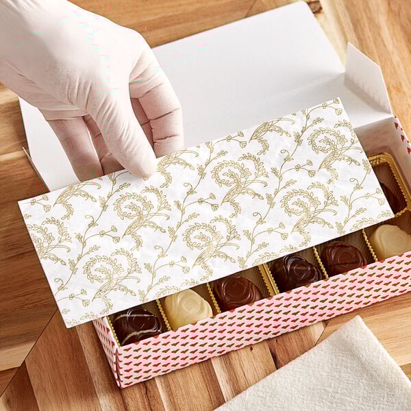 A gloved hand placing a white candy box pad with gold floral pattern in a box of chocolates.