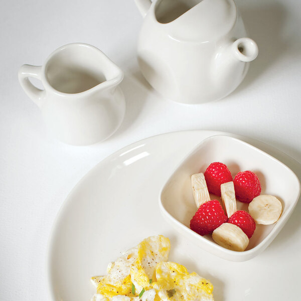 A white pitcher with a handle on a table with a plate of fruit and a bowl of cereal.