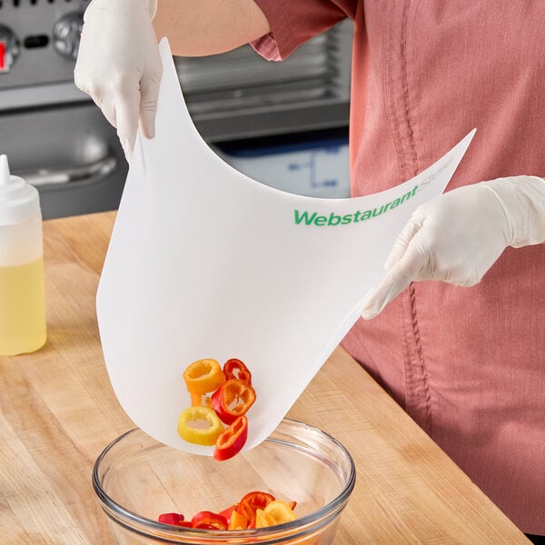 A flexible white cutting board mat with the WebstaurantStore logo being used to transfer sliced peppers into a glass bowl.