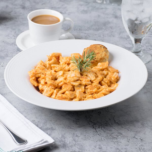 A white porcelain pasta bowl filled with pasta and a piece of bread on a table with a cup of coffee.