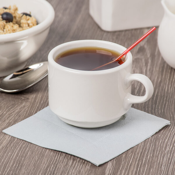 A Libbey ivory porcelain tea cup with a spoon in it on a table with a bowl of oatmeal.