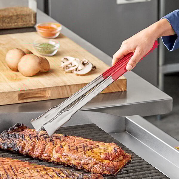 A person using Vollrath VersaGrip tongs with red handles to grill meat.