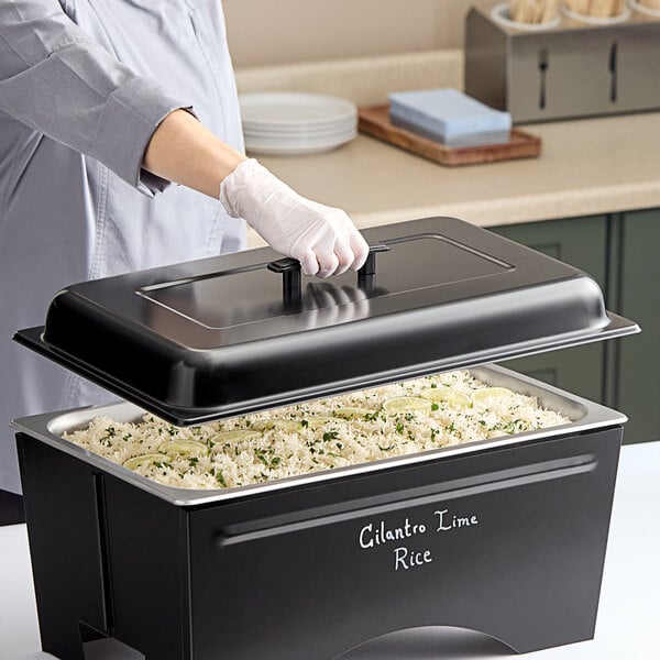 A person wearing gloves holding a black chalkboard lid over a large casserole dish.