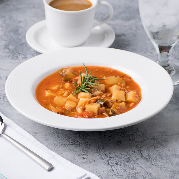 A white porcelain soup bowl filled with soup next to a spoon and a cup of coffee.