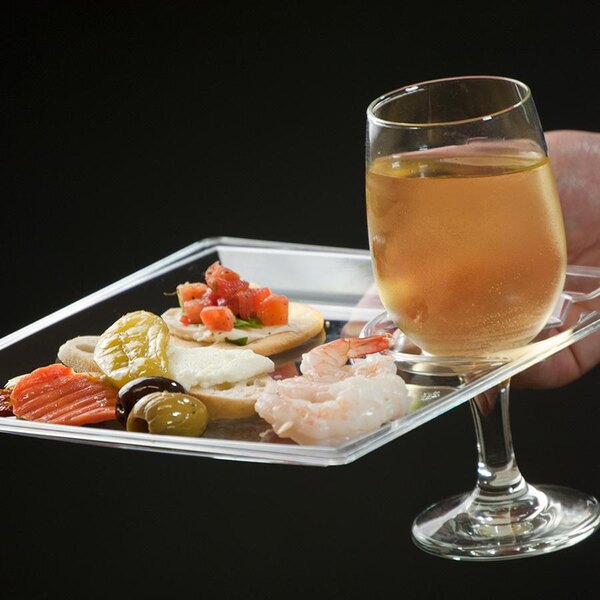 A hand holding a WNA Comet clear square plastic cocktail plate with food.