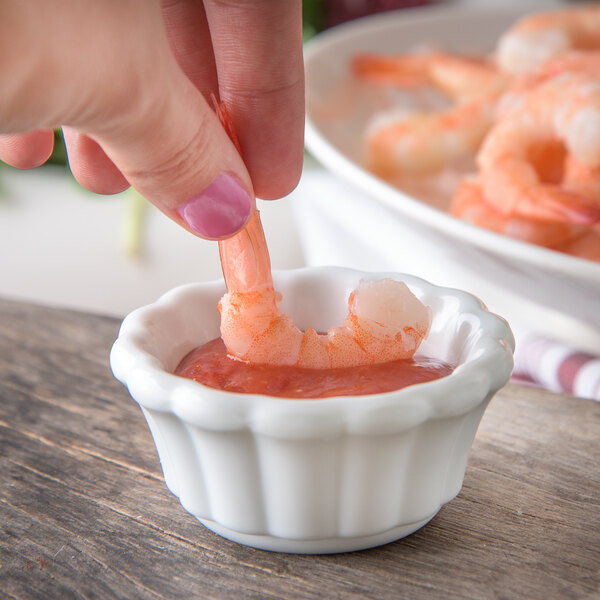 A hand dipping a shrimp into a white Carlisle ramekin of sauce.