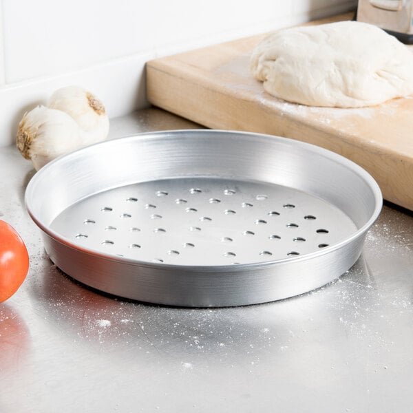 An American Metalcraft silver aluminum pizza pan with holes on it, next to dough and a tomato.