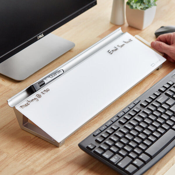 A person using a Quartet white frameless glass dry erase desktop pad on a wooden desk next to a computer.