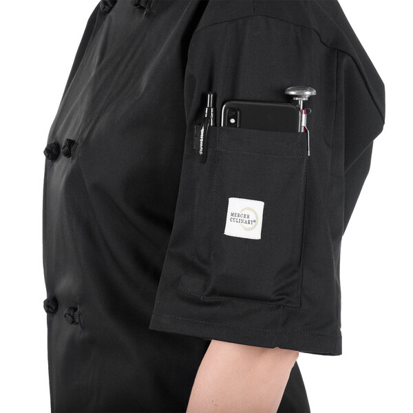 A woman wearing a black Mercer Culinary chef jacket with a phone and pen in the pocket.