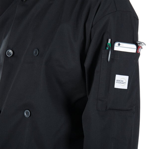 A person wearing a Mercer Culinary Millennia black long sleeve chef jacket with a phone and pen in the pocket.