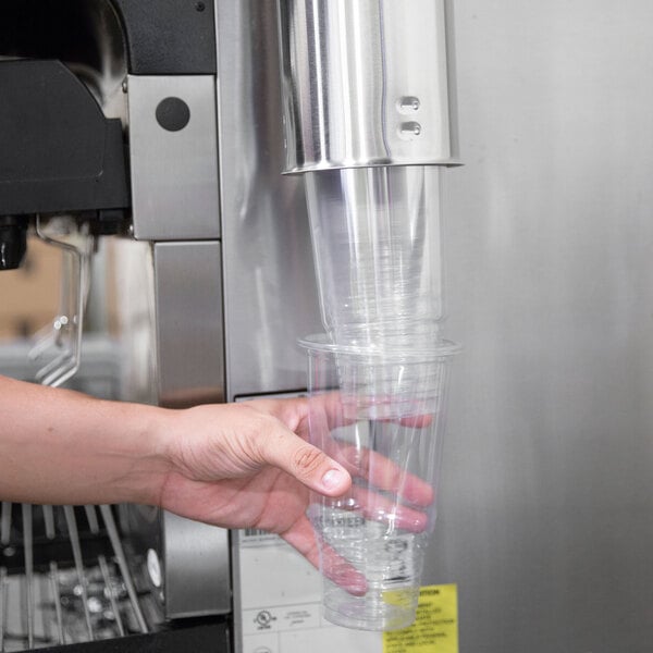 A hand pressing a plastic cup into a Vollrath cup dispenser on a hospital cafeteria counter.