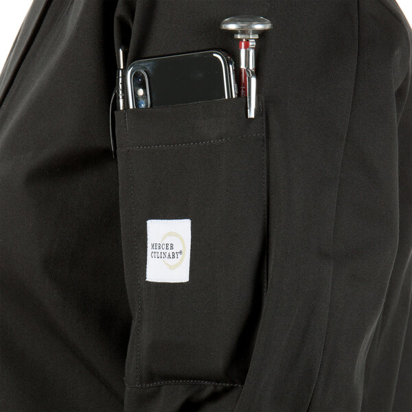 A person wearing a Mercer Culinary Millennia black chef coat with a phone and pen in the pocket.
