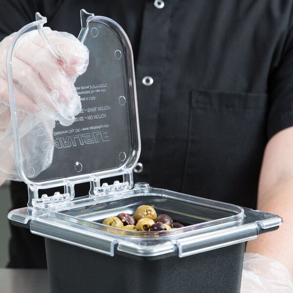 A person wearing a plastic glove using a Carlisle clear plastic food pan lid with a notch to cover a container of olives.