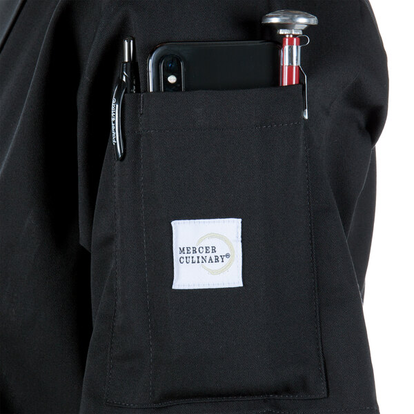 A person wearing a Mercer Culinary Millennia black chef coat with a phone and pen in the pocket.