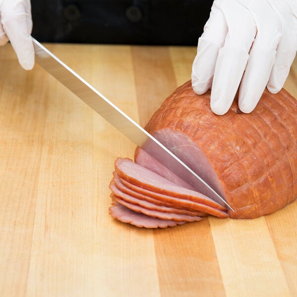 A person using a Mercer Culinary Renaissance forged carving knife to slice ham on a cutting board.