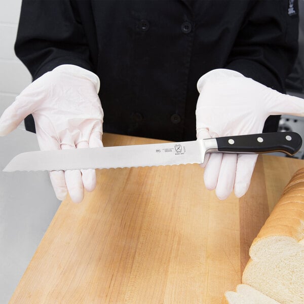 A person in white gloves using a Mercer Culinary Renaissance bread knife to slice a loaf of bread.