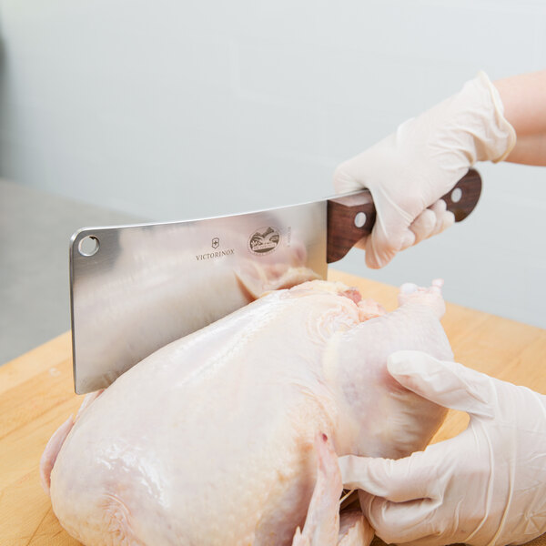 A person in gloves using a Victorinox cleaver to cut a raw chicken on a cutting board.