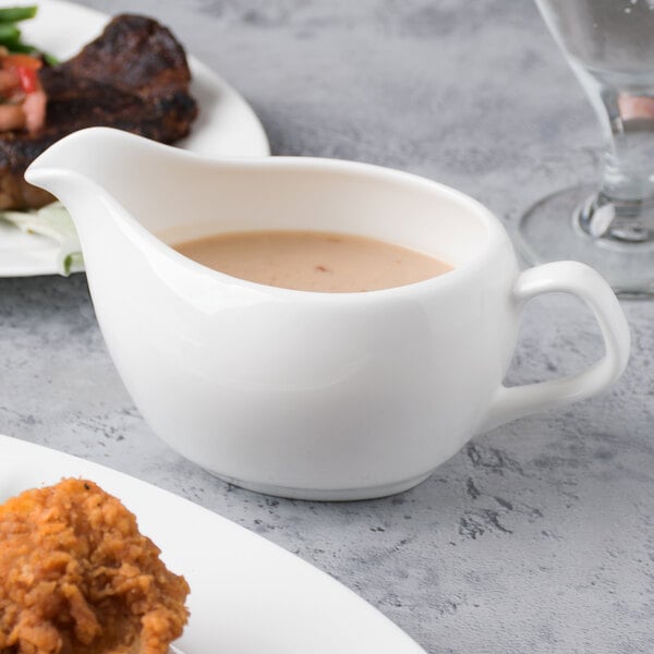 A white gravy boat with sauce on a plate next to steak and vegetables.