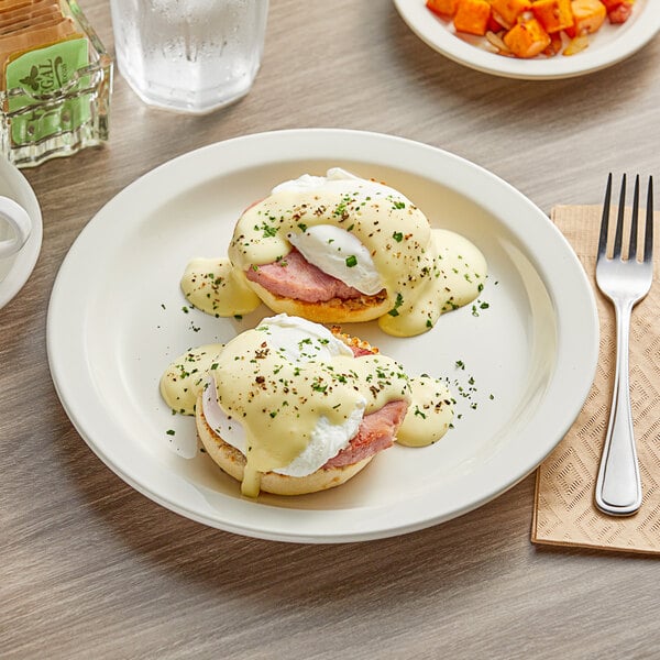 An Acopa ivory stoneware plate with two eggs benedict, ham, and potatoes on a table.