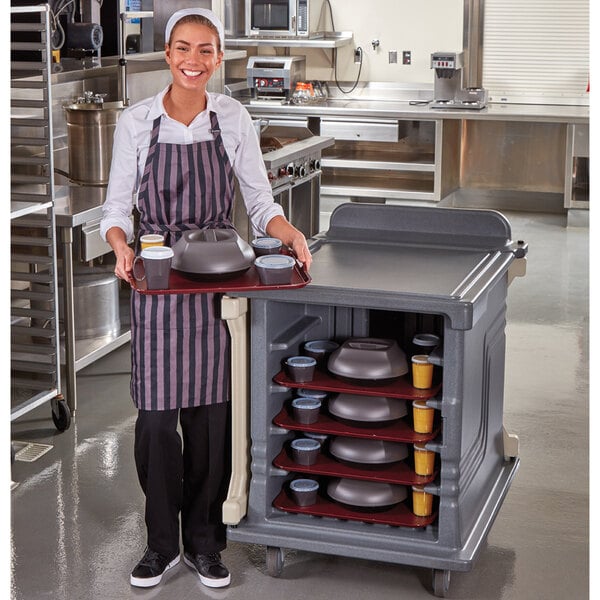 A woman in an apron holding a Cambro meal delivery tray with food.