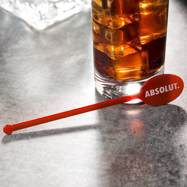 A glass of liquid with ice and a red oval stirrer with the word "absolut" on it.