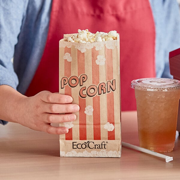 A hand holding a Bagcraft EcoCraft popcorn bag next to a plastic cup of drink.